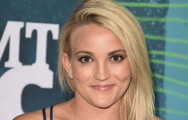 A person with blond hair smiling at an event, wearing a dark blue top against a blue backdrop.