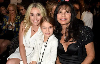 Three women sitting closely together, smiling at the camera, dressed in white and black outfits at an indoor event.