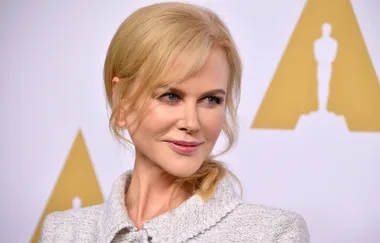 A woman with blonde hair smiling at an event, standing in front of an Oscar statue backdrop.