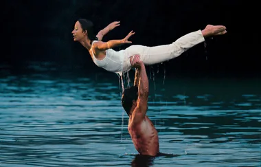 A dancer is lifted in a graceful pose over a lake by a partner, both in white clothing, resembling a scene from "Dirty Dancing."