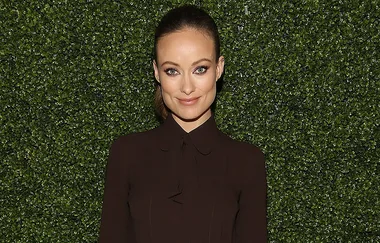 Smiling person in front of a green hedge wall, wearing a dark buttoned-up shirt.