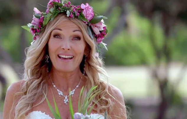 Blonde woman with a flower crown and necklace, smiling outdoors.