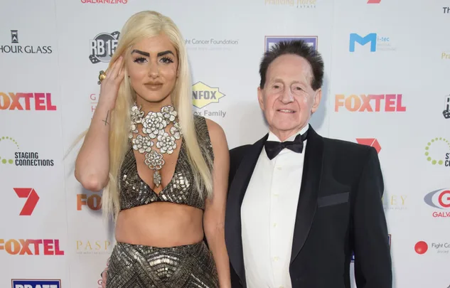 Two people posing on a red carpet event, one in a metallic outfit, the other in a tuxedo, with logos in the background.