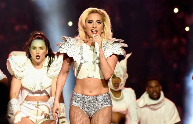 Singer in a dazzling outfit performs energetically on stage with backup dancers in matching attire.