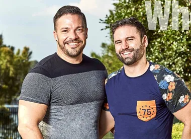 Two men smiling outdoors with trees in the background, one wearing a black and grey shirt, the other a floral sleeve shirt.