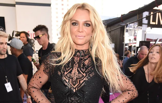 I don't know who this person is, but they're posing in a black, lace dress at an event with people and cameras around.