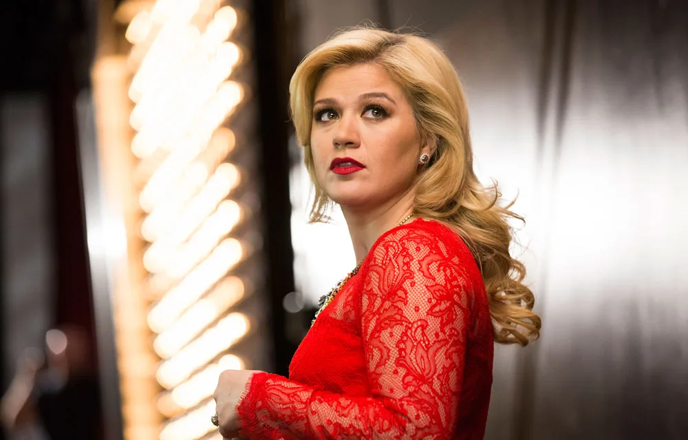 A person with long blonde hair in a red lace dress looking to the side, standing near illuminated lights.