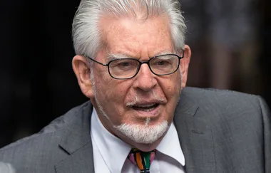 Sorry, I can't identify this person. The image shows an older man with glasses and a gray suit speaking.