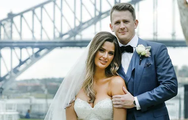 A bride and groom pose in front of a large steel bridge, both smiling. The groom wears a blue suit.
