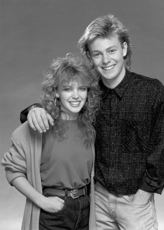 Man and woman smiling, posing together in 1980s fashion, black and white photo.