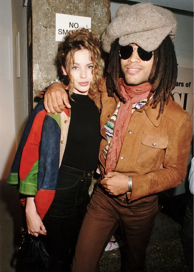 A person with long hair and sunglasses in a brown outfit poses beside another person in colorful attire.
