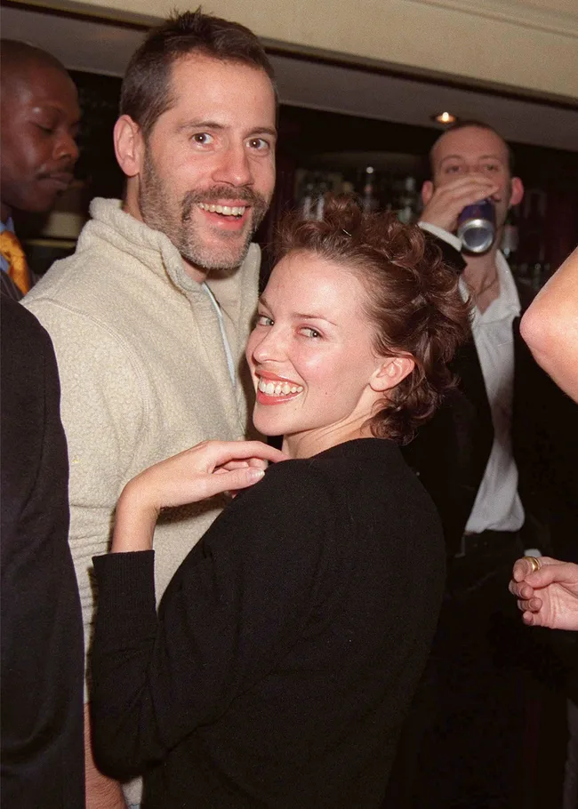 Two people smiling at a gathering with others in the background, one drinking from a can.