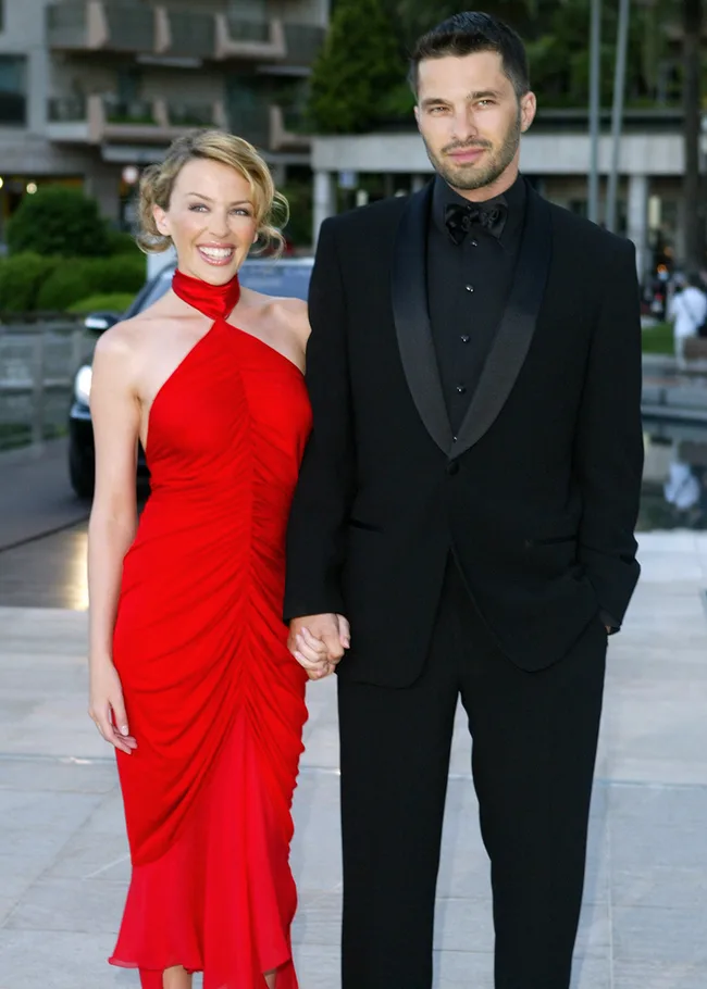 Woman in a red dress and man in a black tuxedo holding hands, outdoors with buildings in the background.
