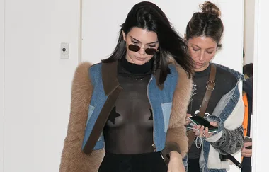 Two people walking indoors; one in sunglasses and a sheer top with star details, the other looking at a device.