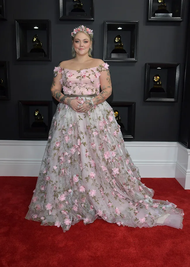 Person in a floral gown with flower headpiece on a red carpet, Grammys backdrop with awards visible.