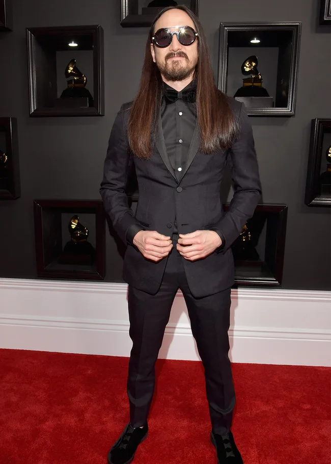 Man with long hair and sunglasses in a black suit on the red carpet at an awards event.