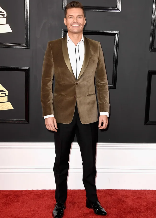 Man in a brown velvet blazer, white shirt, and black trousers on a red carpet at a formal event.
