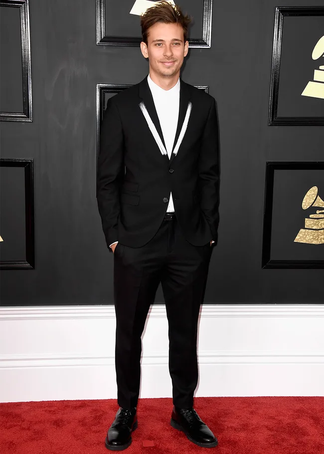 Young man in a black suit and white shirt stands on a red carpet at an event, with framed Grammy symbols in the background.