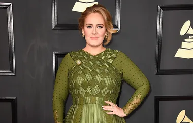 A person in an elegant green dress poses at the Grammy Awards in front of a black background with framed logos.