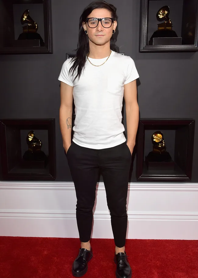 A person in a white shirt and black pants on a red carpet with Grammy awards in the background.