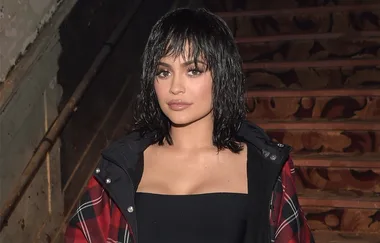 Young woman with dark hair wearing a plaid jacket stands on a staircase.