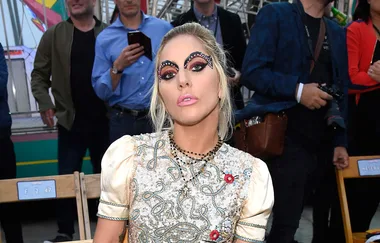 A person with elaborate eye makeup and embroidered top sits among a crowd.