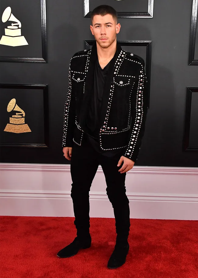 Man in a studded black jacket on a red carpet, posing in front of Grammy logos.