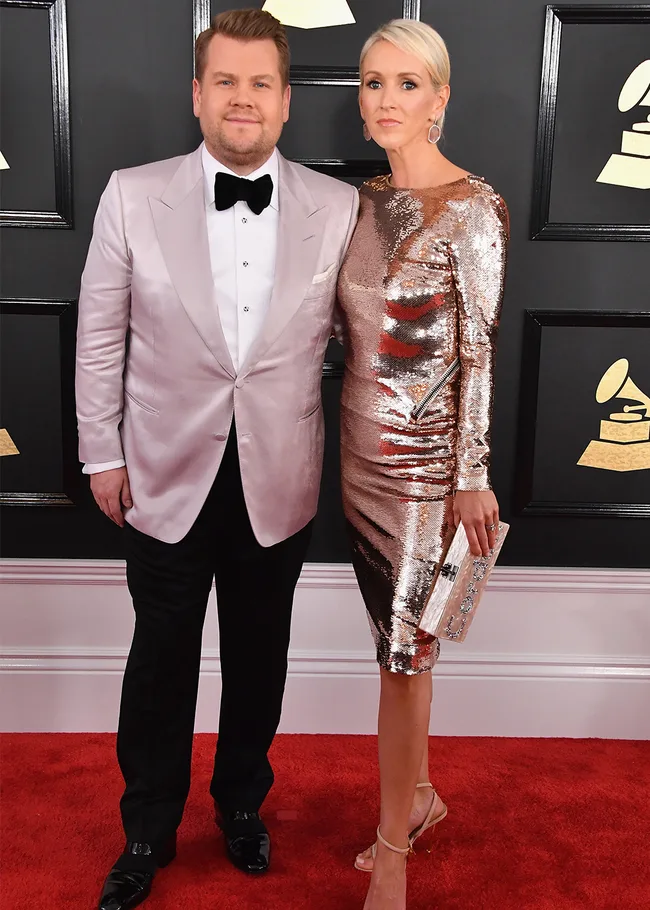 Man in pink tuxedo and woman in silver dress pose on a red carpet at an awards event.