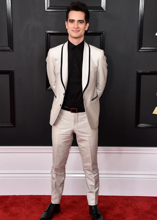 A person in a white suit with black details and a black shirt stands on a red carpet against a dark background.