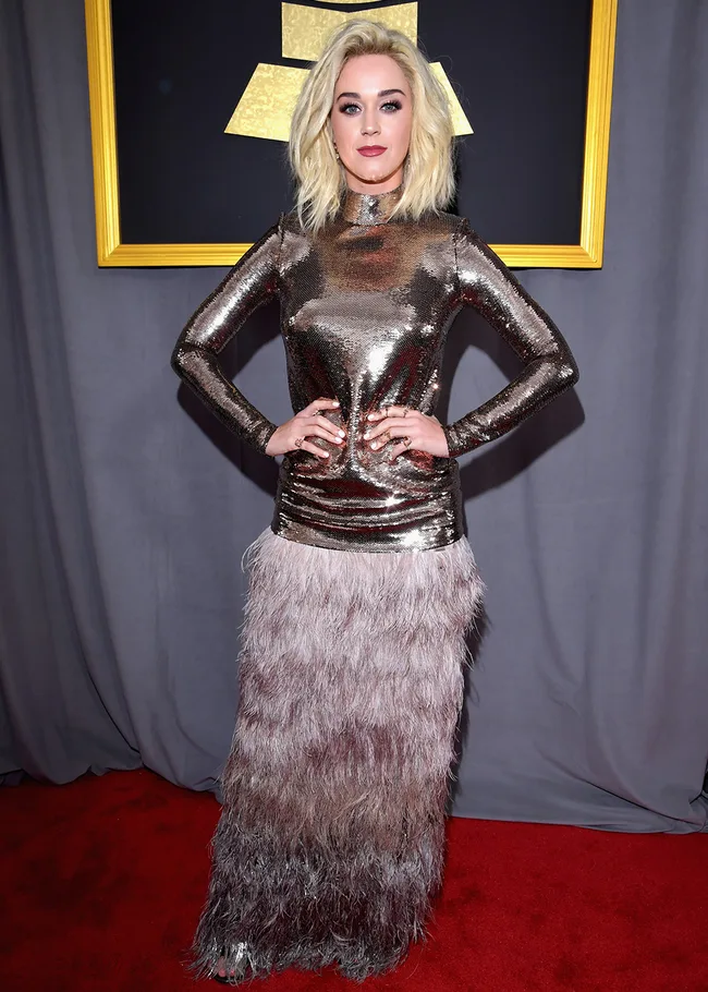 Woman in a metallic, sequined gown with feathered skirt poses on a red carpet in front of a framed award logo.