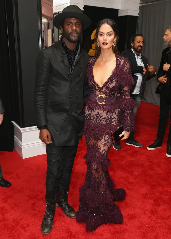 A man in a black suit and hat stands with a woman in a lace burgundy gown on a red carpet event.