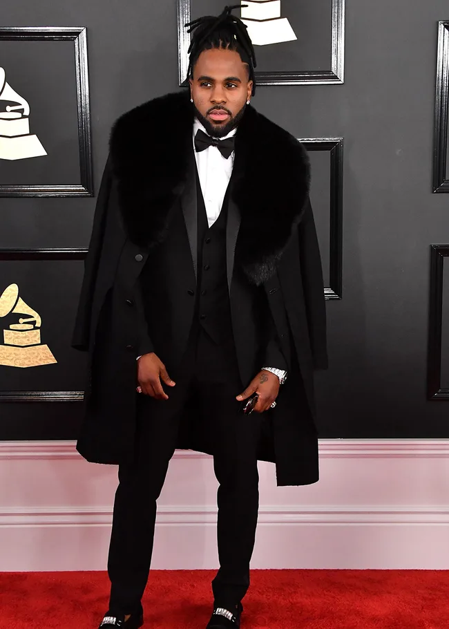 Man in a black tuxedo with fur stole poses on red carpet at an awards event.
