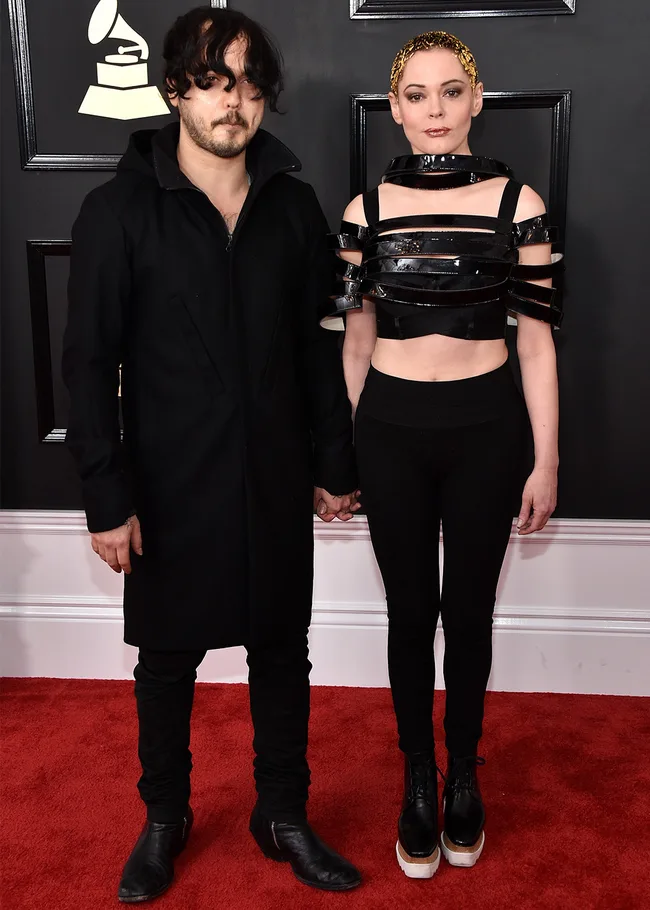 A man in black outfit and a woman in a black strapped top and pants stand on the red carpet holding hands.