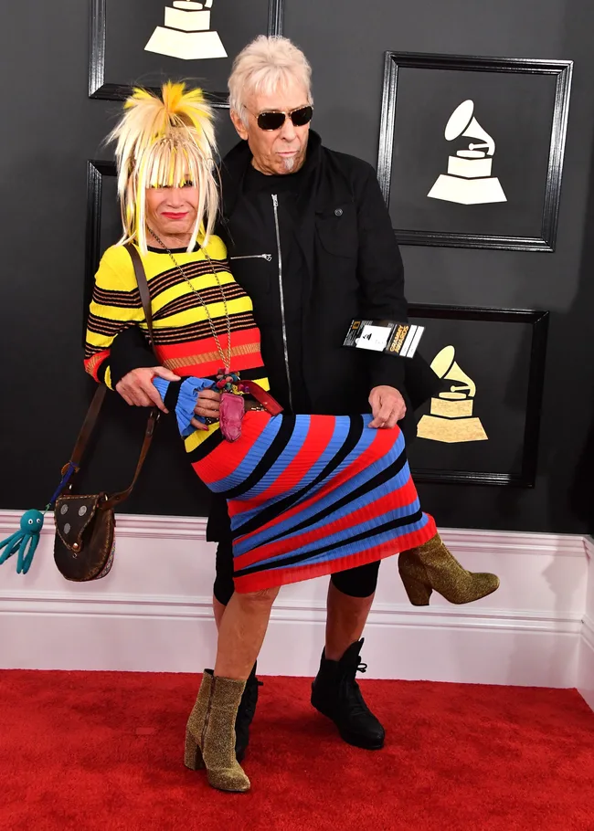 Two people pose on a red carpet, one holding the other; colorful striped dress, sunglasses, and Grammy backdrop visible.