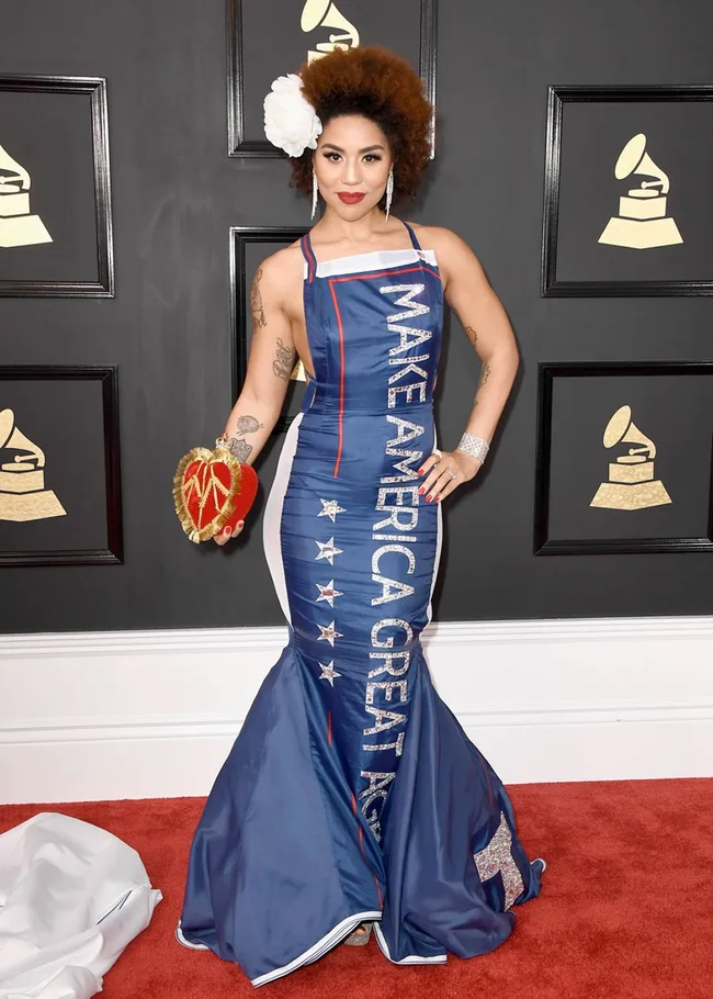 A person in a blue dress with "MAKE AMERICA GREAT AGAIN" text, at a Grammy Awards event, holding a heart-shaped clutch.