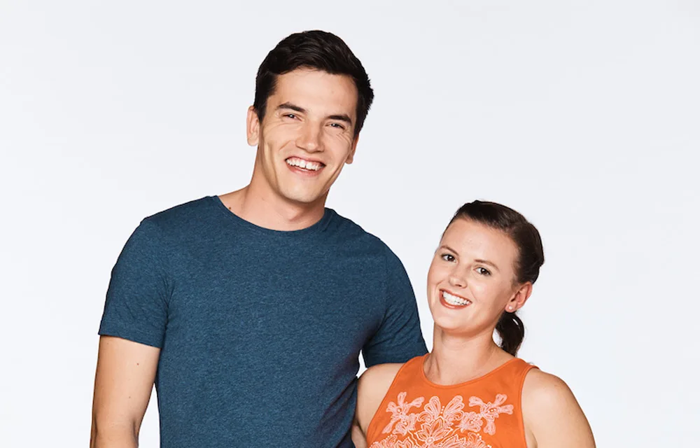 A man in a blue shirt stands smiling next to a woman in an orange top against a plain background.