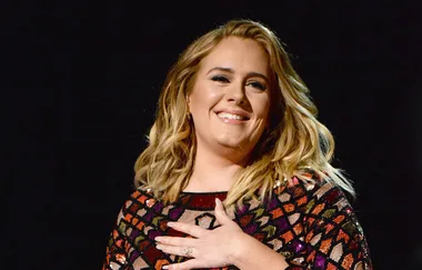 A person smiling, with wavy blonde hair, wearing a colorful patterned top against a dark background.