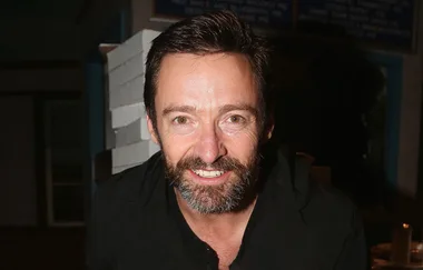 A person with dark hair and a beard smiling in a dimly lit room, wearing a dark shirt.