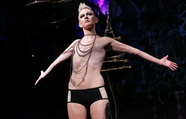 Person on stage wearing chains, arms outstretched, under dramatic lighting.