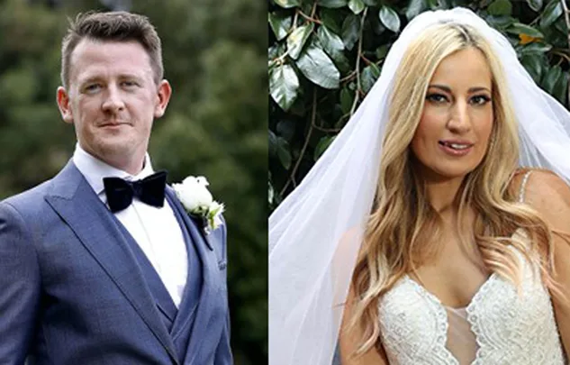 Groom in blue suit with bow tie and bride in white lace dress with veil, posing outdoors.