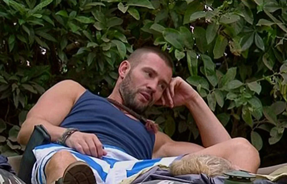 A man in a sleeveless shirt and shorts reclines outdoors, surrounded by greenery.