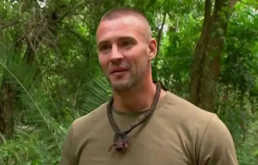 Man in green shirt with necklace, standing in lush forest setting.