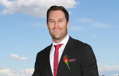 A man in a suit with a red tie and rose lapel stands outside against a blue sky background.