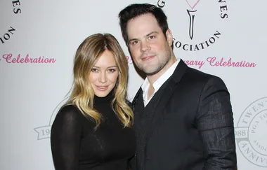 A woman in a black dress and a man in a suit pose together at an event with a white and pink logo backdrop.