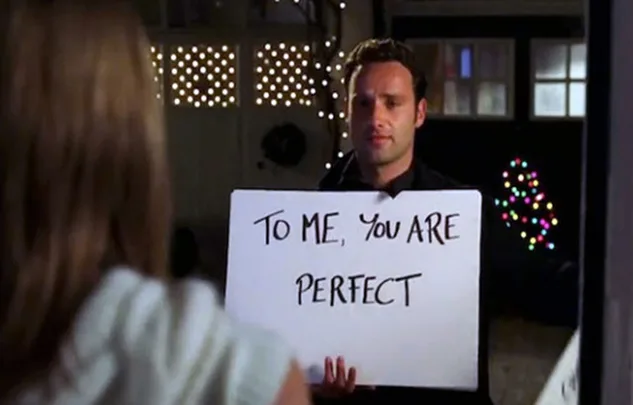 Man holding a sign that says "To me, you are perfect" outside a lit home during a scene from the film Love Actually.