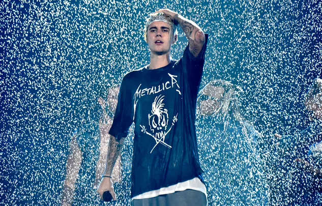 Singer in a wet Metallica shirt holding a microphone, surrounded by falling water on stage.