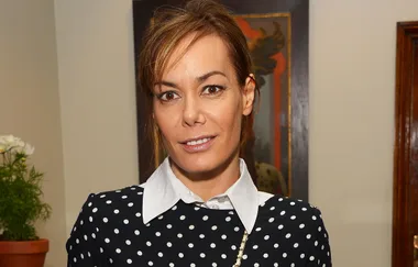 A person with brown hair in a polka-dot top stands indoors, smiling.