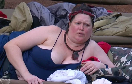 Woman in navy swimsuit lying on a camp bed, surrounded by sleeping bags and clothing, with a concerned expression.