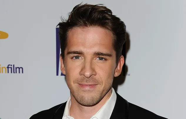 Portrait of a man with brown hair, wearing a black suit and white shirt, against a light background with a logo.