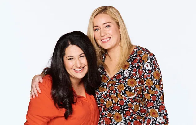 Two women smiling, one with dark hair in orange, the other with blonde hair in a floral top, posing together.